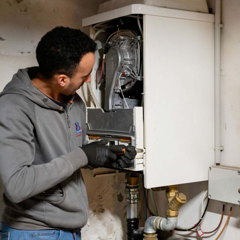 Un tecnico, indossando guanti e una felpa grigia, sta lavorando su una caldaia aperta. Utilizza un cacciavite per effettuare delle regolazioni o per rimontare una parte del pannello. L'interno della caldaia mostra componenti meccanici e tubature, suggerendo un intervento di manutenzione o riparazione. Il tecnico appare concentrato, operando con attenzione in un ambiente che sembra essere un locale tecnico o una sala caldaie.