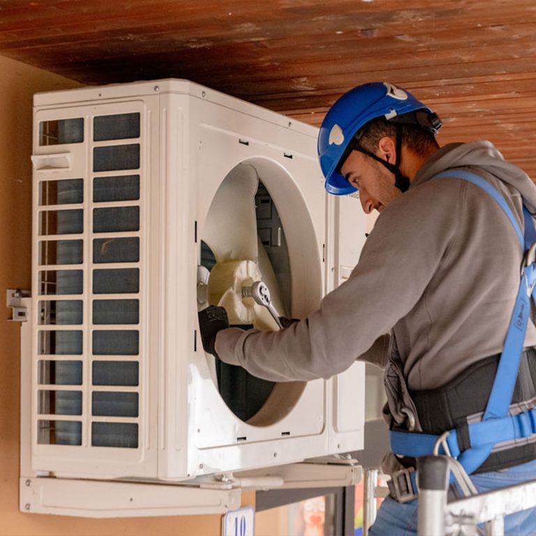 Un tecnico indossa un casco di sicurezza blu e un'imbracatura mentre lavora su un'unità esterna di climatizzazione. È intento a fare manutenzione o riparare la ventola all'interno dell'unità. Il tecnico indossa guanti e una felpa grigia, mostrando attenzione alla sicurezza e alla protezione. L'unità è montata su una parete esterna, e il tecnico sembra concentrato nel suo lavoro, che richiede precisione e competenza. L'immagine trasmette un ambiente di lavoro sicuro e professionale.